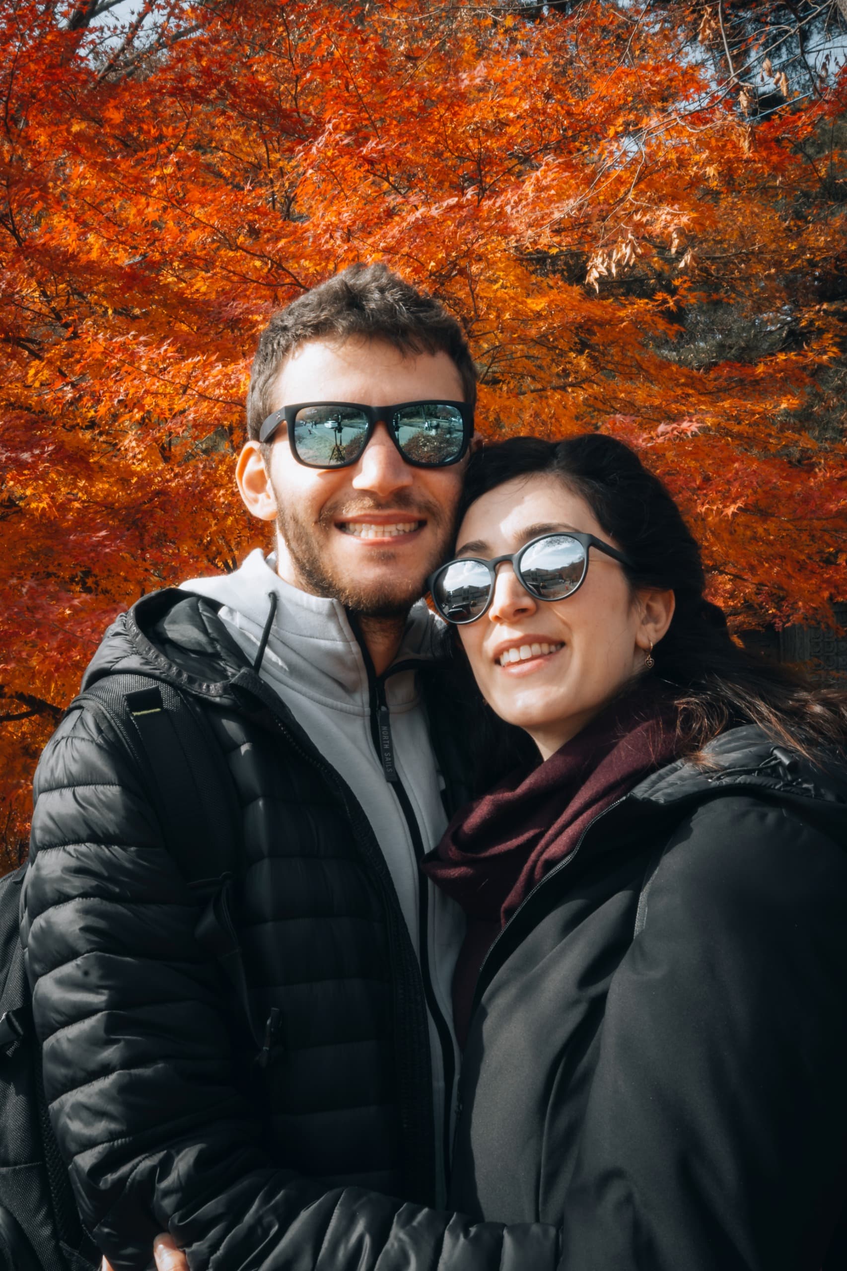 Marco e Martina abbracciati e sorridenti davanti a un acero dai colori autunnali.